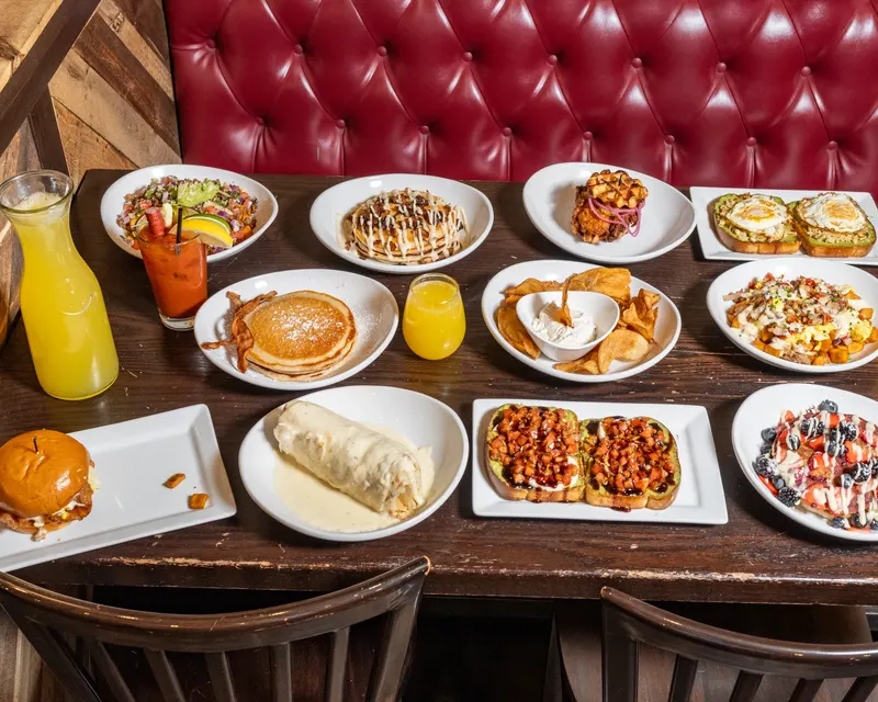 A table at Oak Barrel covered in brunch plates, among them pancakes, a burrito and a burger, with a pitcher of orange juice