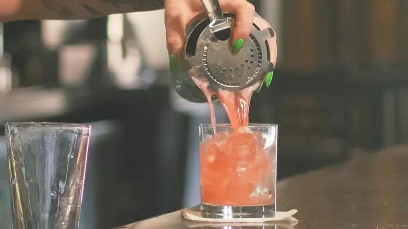 A bartender straining a pink cocktail from a shaker into a glass on the bar